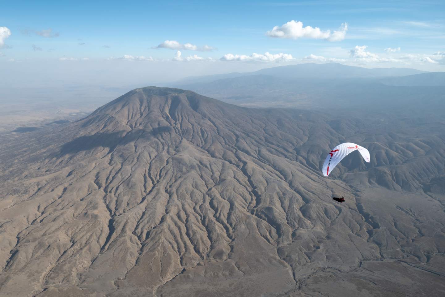 Pilote de hike & fly en montée vers le décollage, sac compact contenant voile et sellette sur le dos, crêtes de montagne en arrière-plan.